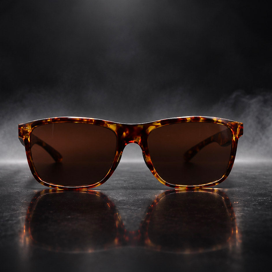 Tortoiseshell square sunglasses with dark brown lenses, displayed on a reflective dark surface with a dramatic smoky background.
