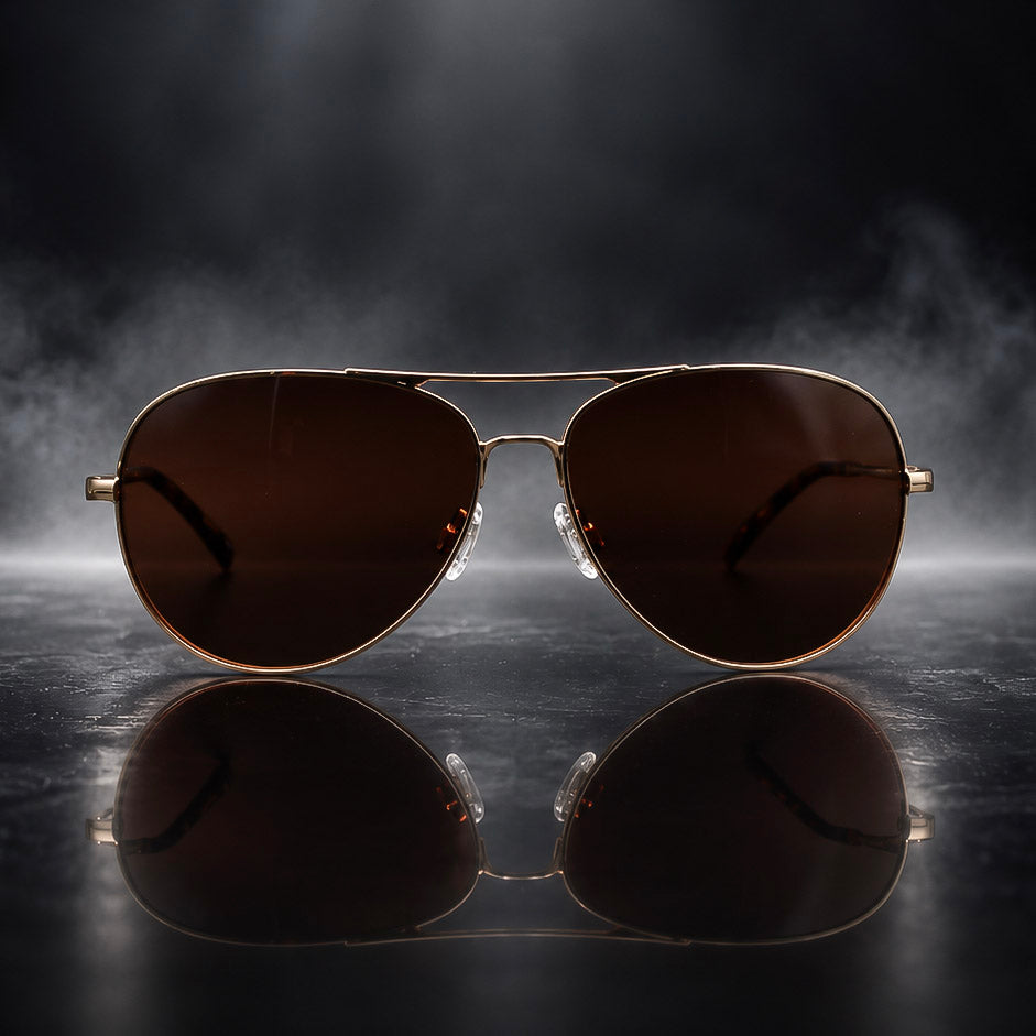 A pair of classic aviator sunglasses with dark brown lenses and a gold metal frame, displayed on a reflective dark surface with a dramatic smoky background.
