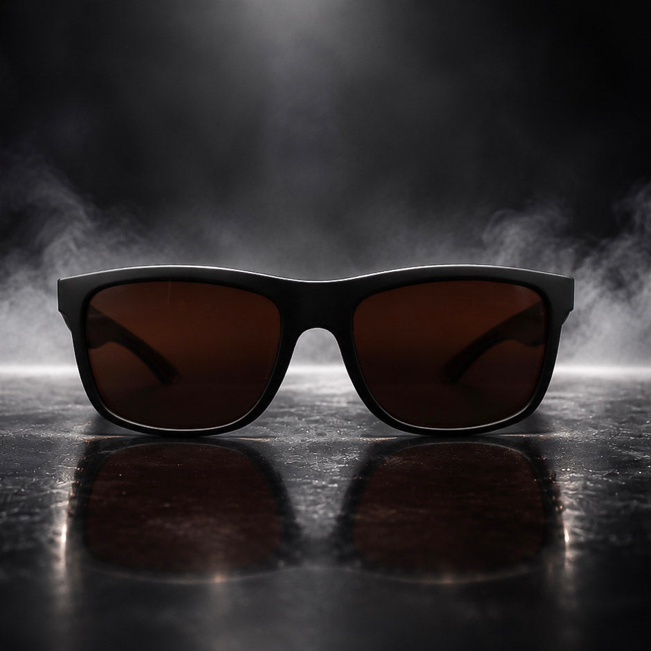 Black square sunglasses with dark brown lenses resting on a reflective dark surface with a smoky background.